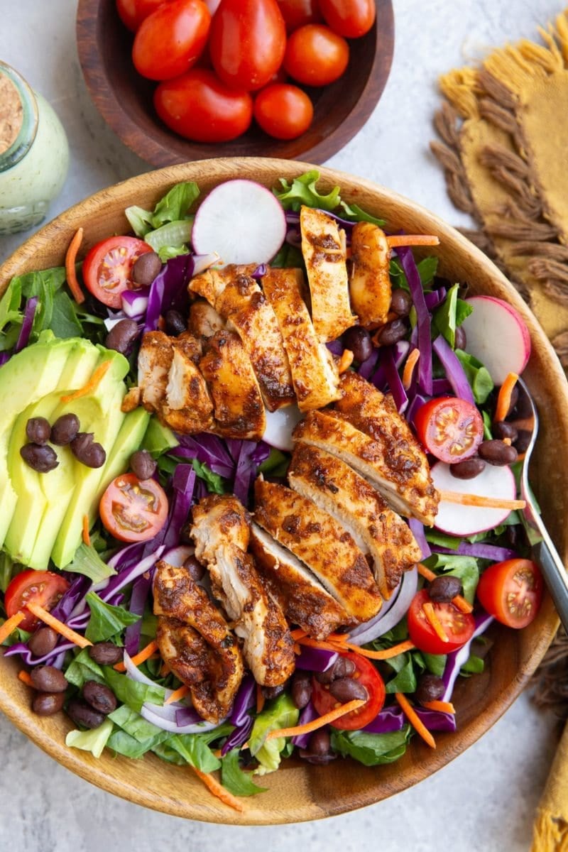 Large bowl of taco salad with fresh spring greens, black beans, sliced avocado, cherry tomatoes, cabbage, and more.