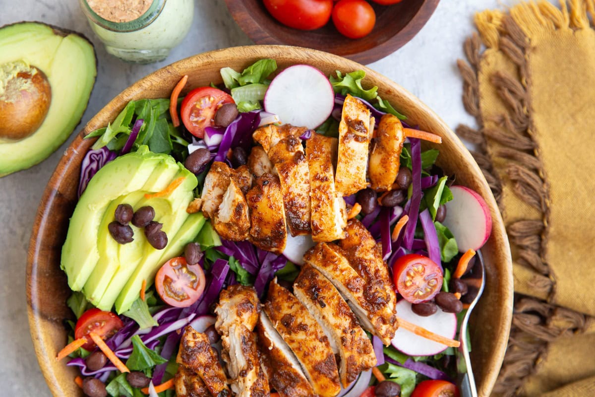 Chicken salad in a wooden bowl with beans, avocado, tomatoes, cabbage, and more. Half of an avocado to the side and a golden napkin.