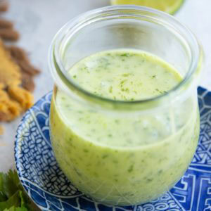 Jar full of cilantro lime dressing on top of a blue patterned plate with fresh limes in the background and fresh cilantro in the foreground.