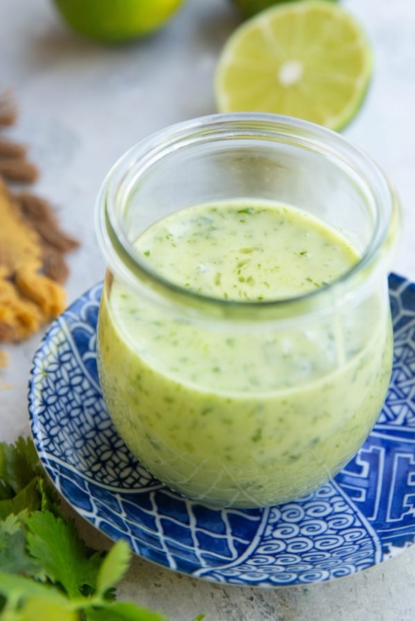 Jar full of cilantro lime dressing on top of a blue patterned plate with fresh limes in the background and fresh cilantro in the foreground.