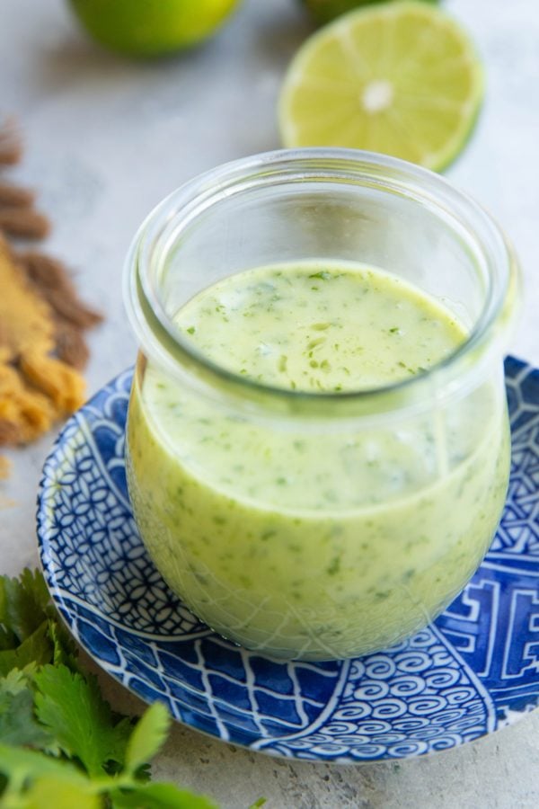 Jar full of cilantro lime dressing on top of a blue patterned plate with fresh limes in the background and fresh cilantro in the foreground.