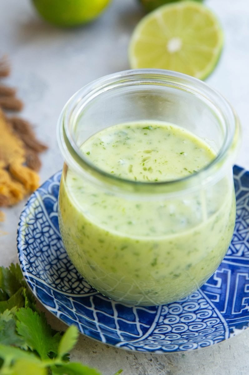 Jar full of cilantro lime dressing on top of a blue patterned plate with fresh limes in the background and fresh cilantro in the foreground.