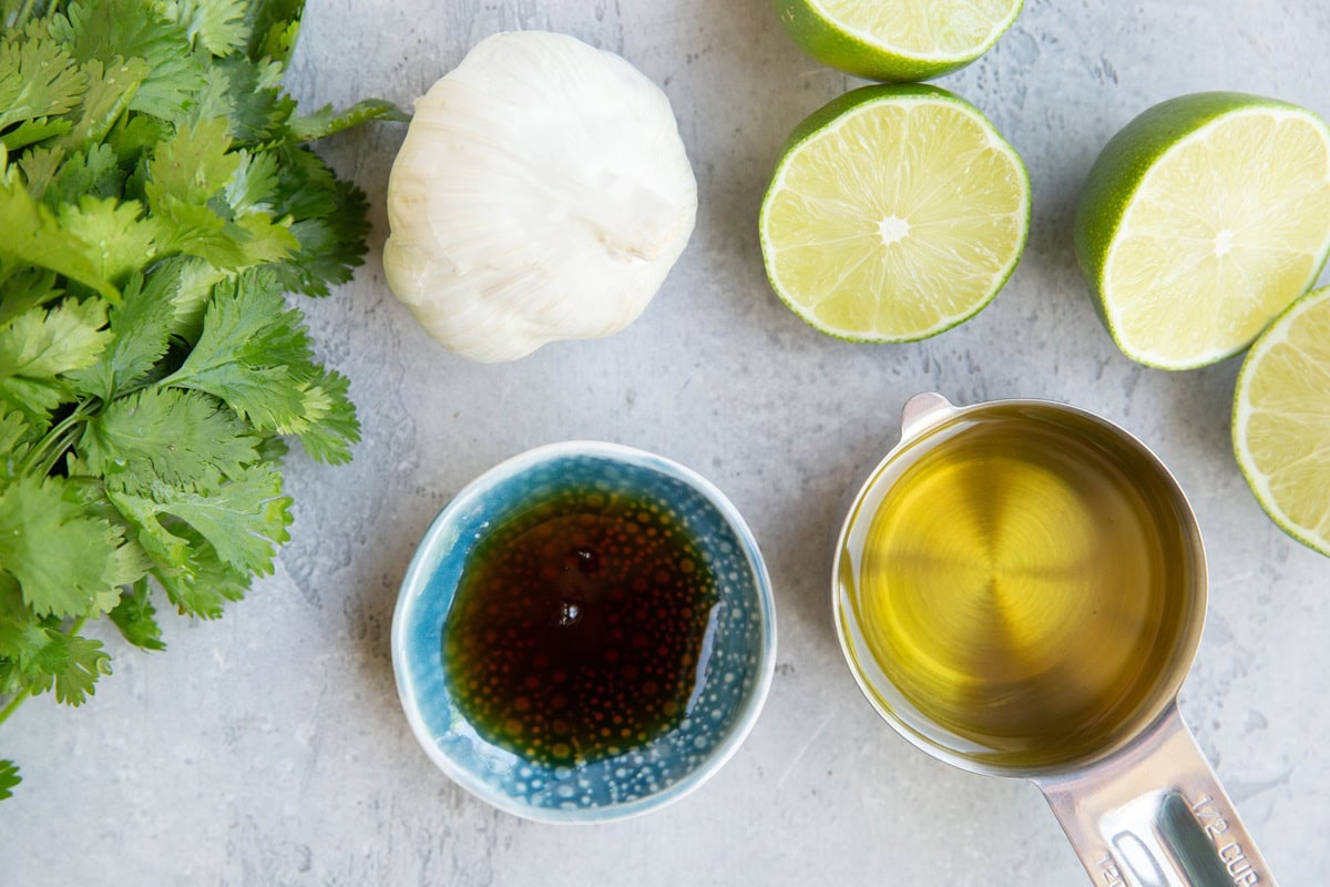 Ingredients for cilantro lime dressing on a flat surface. Fresh cilantro, oil, fresh limes, fresh garlic, and pure maple syrup.