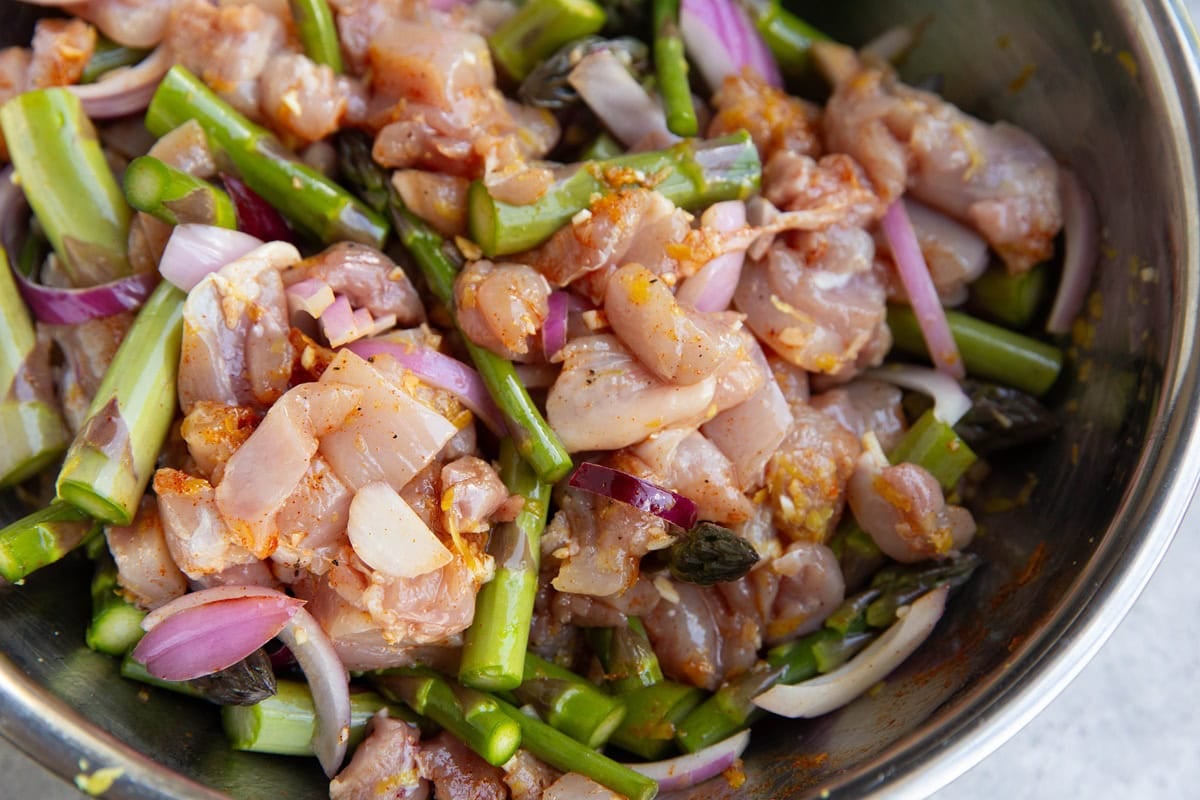 Stainless steel bowl full of chicken, asparagus, and onion tossed in oil and seasonings.