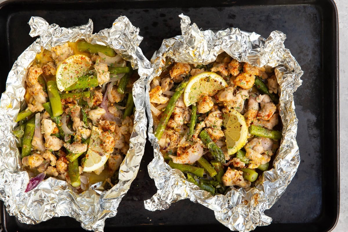 Baking sheet with two packets of aluminum foil with chicken and asparagus.
