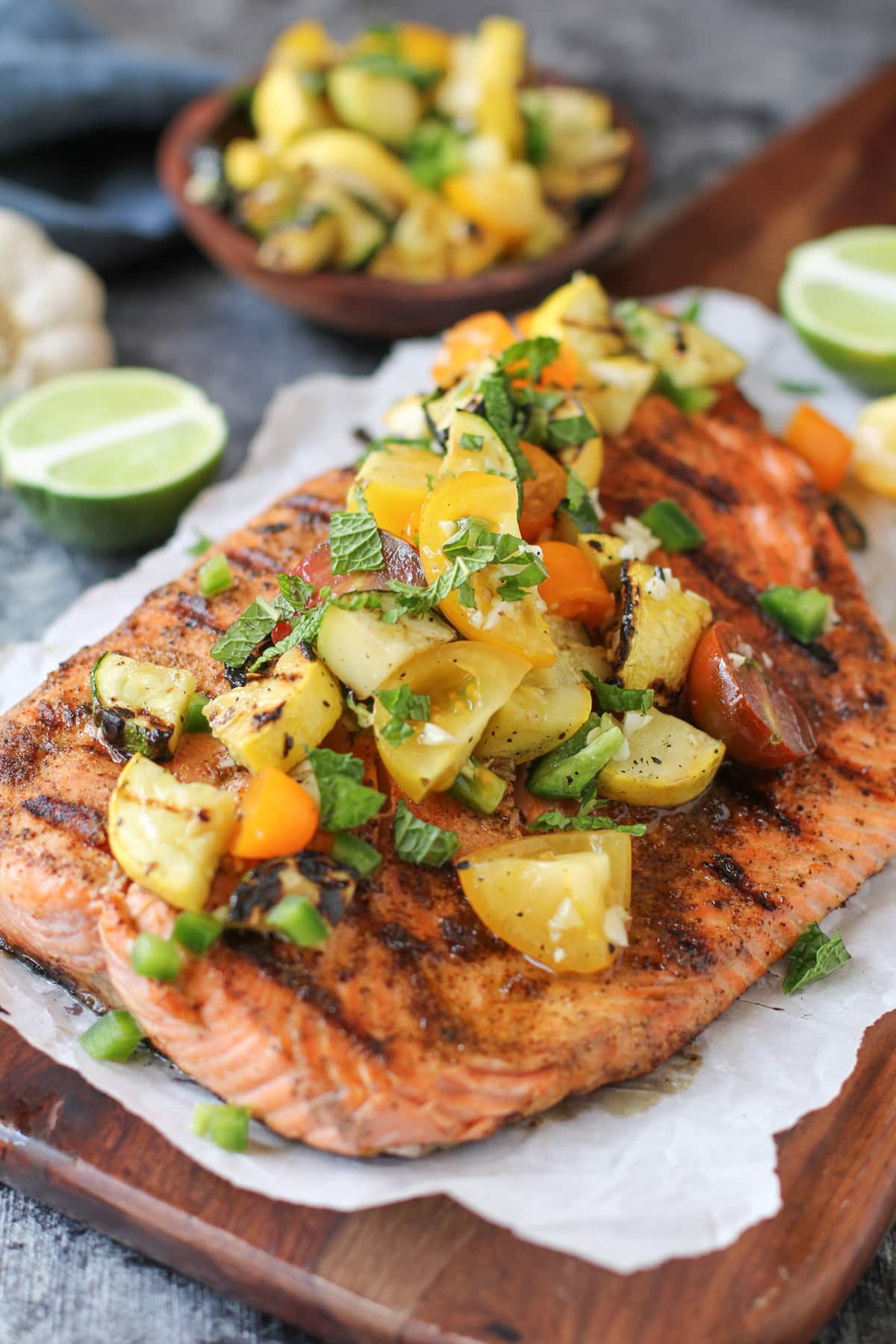 Large grilled salmon filet on a cutting board with fresh chopped tomatoes, grilled zucchini, mint, and green onions sprinkled on top.