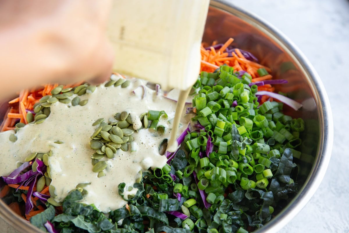 Large bowl full of kale slaw ingredients with dressing being poured in.