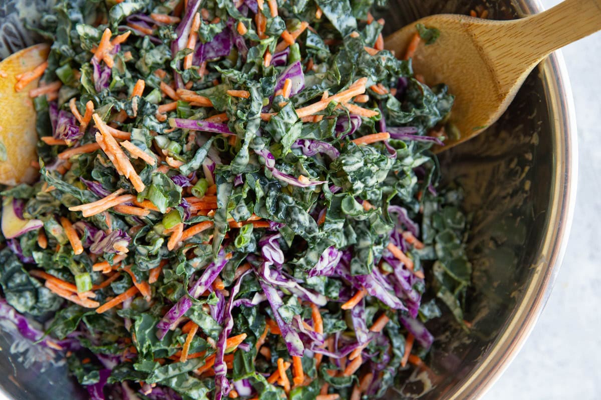 Kale slaw in a stainless steel bowl with wooden spoons inside. Ready to serve.