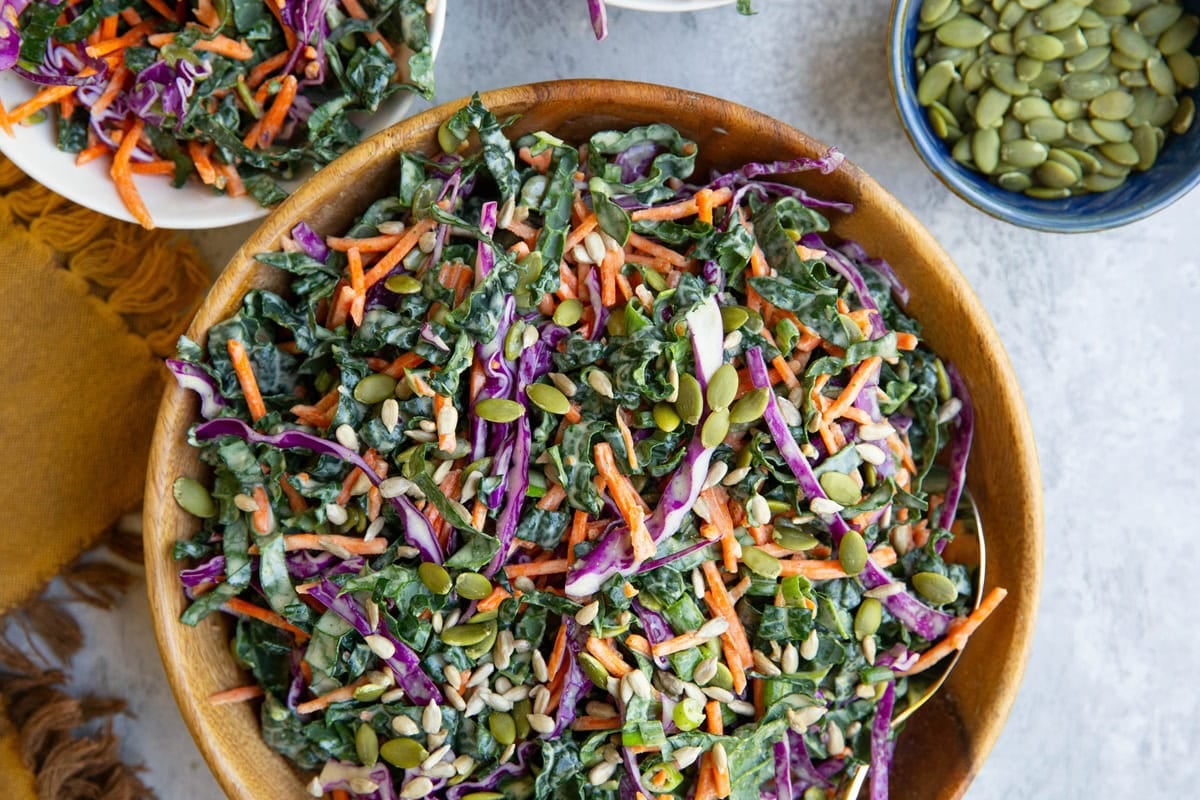 Wooden bowl full of kale slaw with carrots, cabbage, pumpkin seeds, and creamy dressing.