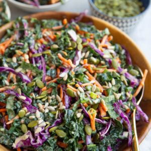 Kale slaw recipe in a wooden bowl with a golden spoon, ready to serve. a bowl in the background with a serving of slaw and a bowl of pumpkin seeds.