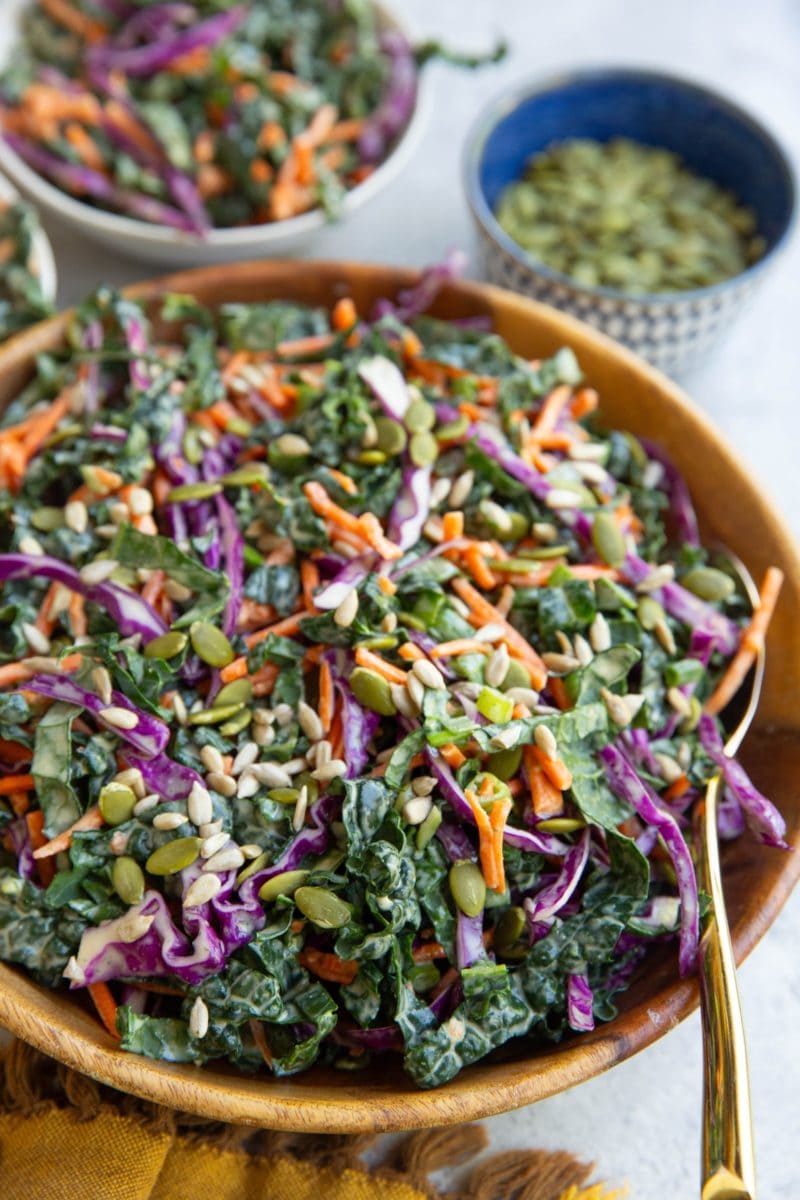 Kale slaw recipe in a wooden bowl with a golden spoon, ready to serve. a bowl in the background with a serving of slaw and a bowl of pumpkin seeds.