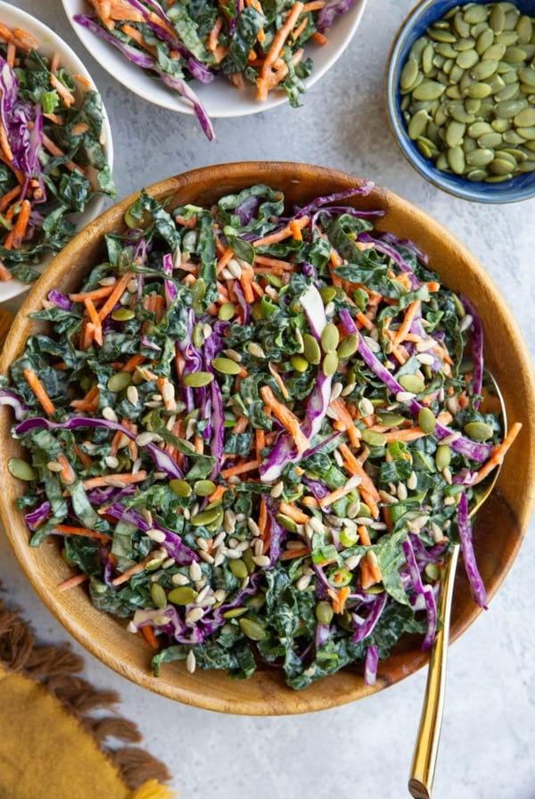 Kale slaw in a wooden bowl and two small bowls with servings of slaw. A small bowl of pumpkin seeds and a napkin to the side.