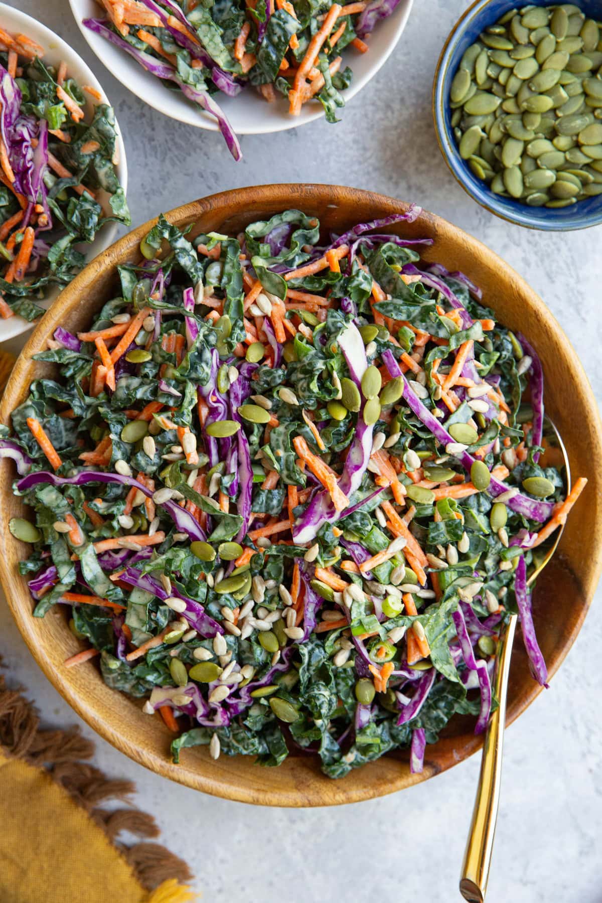 Kale slaw in a wooden bowl and two small bowls with servings of slaw. A small bowl of pumpkin seeds and a napkin to the side.