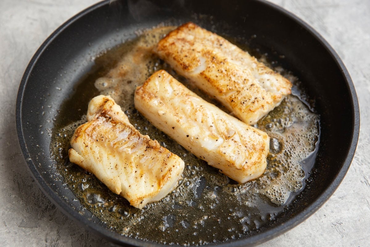 Three cod fillets cooking in a nonstick skillet in butter and oil with a golden brown sear on the outside.