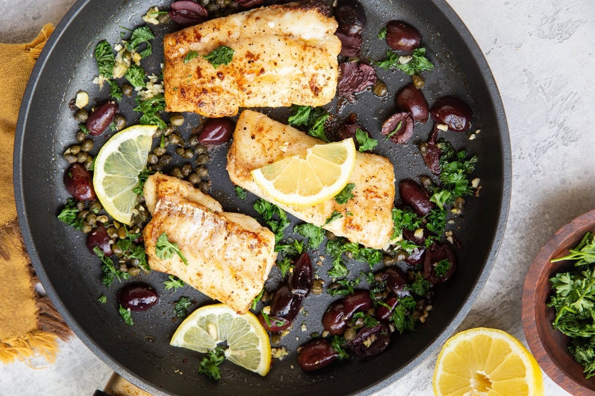 Three pan-fried cod filets in a skillet with fresh lemons, kalamata olives, capers, and parsley. A bowl of fresh parsley to the side.
