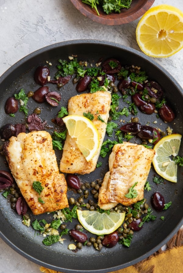 Three seared cod filets in a nonstick frying pan with lemon slices, capers and olives.