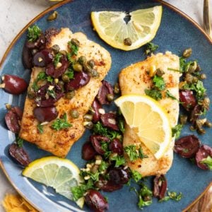 Two cod fillets on a blue plate sprinkled with olives, capers, parsley and lemon slices. Ready to serve.