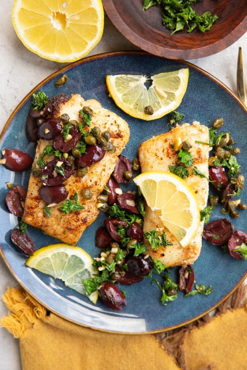 Two cod fillets on a blue plate sprinkled with olives, capers, parsley and lemon slices. Ready to serve.