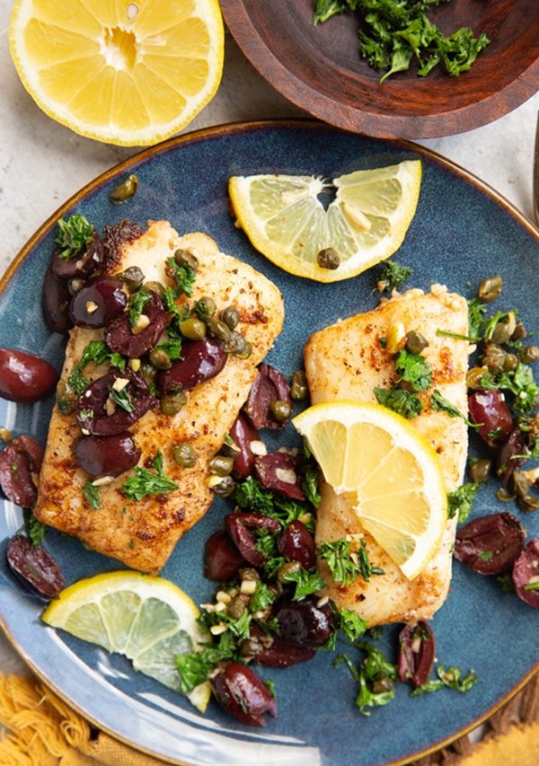 Plate with pan-fried cod with olives and capers on top.