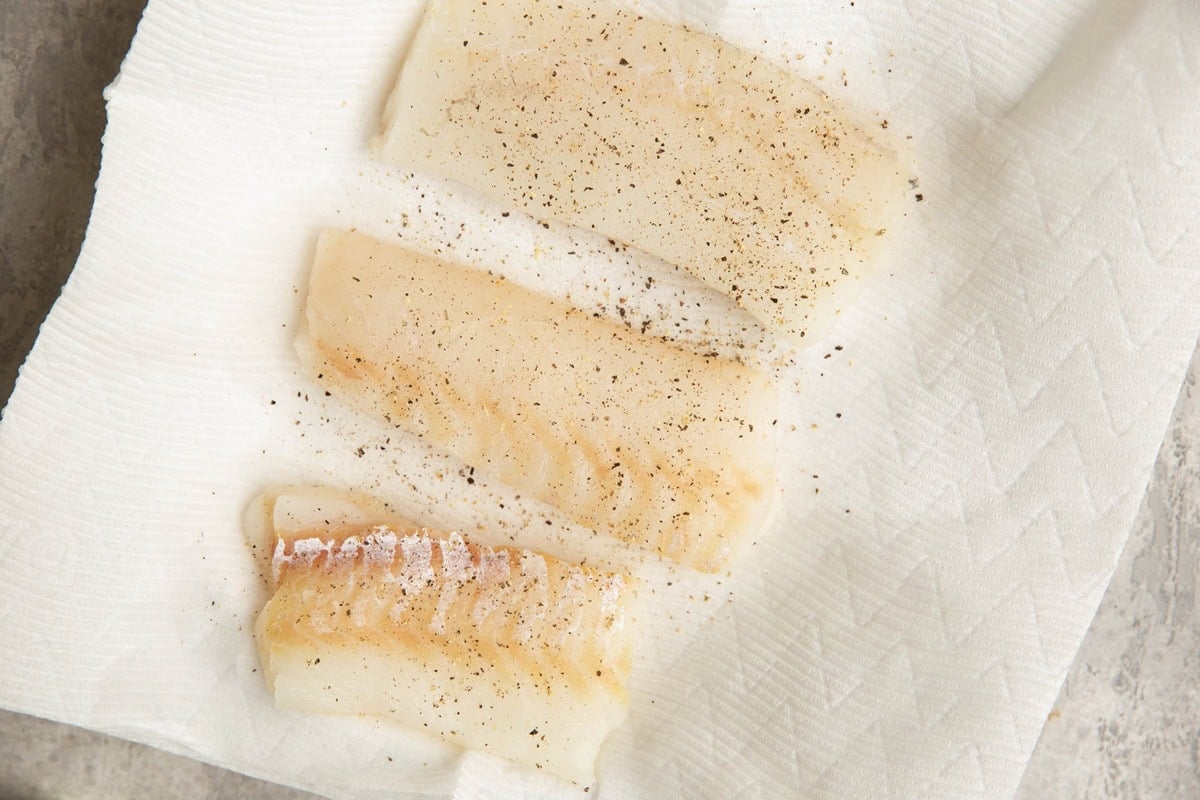 Three fresh cod filets on a plate lined with paper towels, sprinkled with salt and pepper.