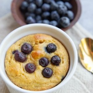Almond flour blueberry muffin in a mug with a bowl of blueberries in the background. A gold spoon to the side, ready to serve.