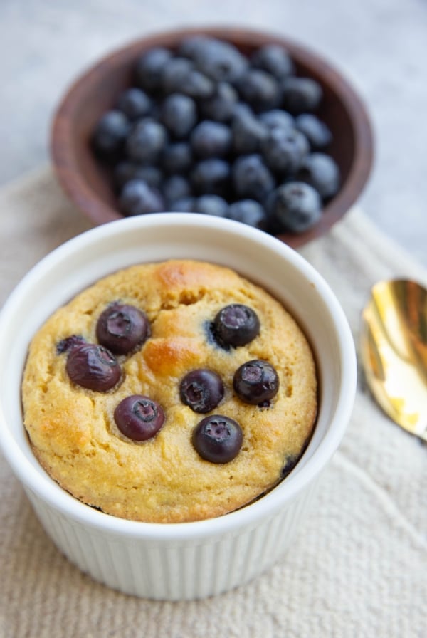 Almond flour blueberry muffin in a mug with a bowl of blueberries in the background. A gold spoon to the side, ready to serve.