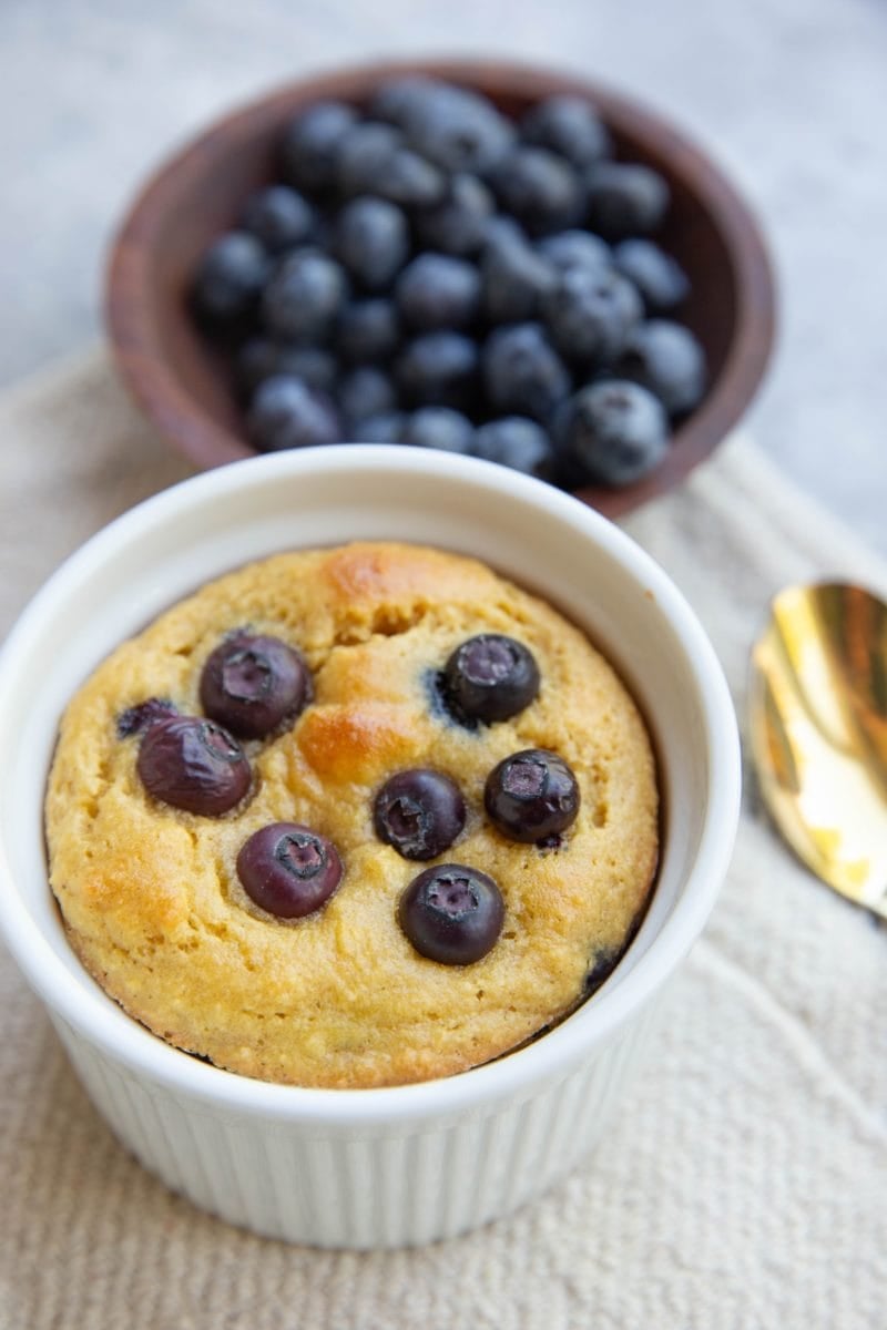 Almond flour blueberry muffin in a mug with a bowl of blueberries in the background. A gold spoon to the side, ready to serve.