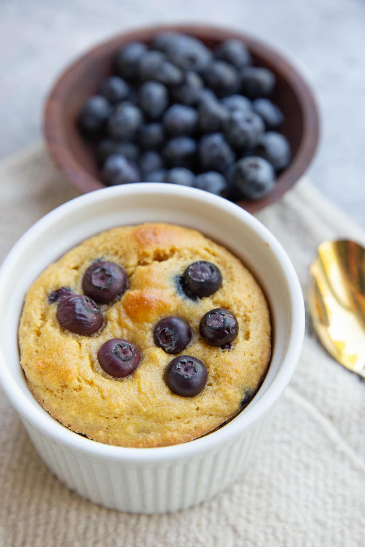 Almond flour blueberry muffin in a mug with a bowl of blueberries in the background. A gold spoon to the side, ready to serve.
