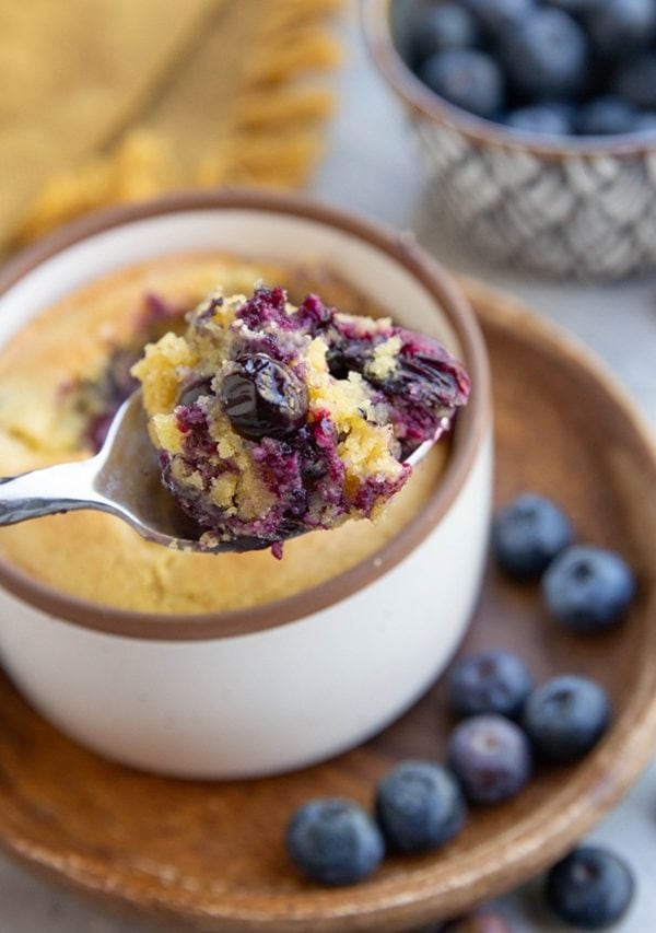 A spoonful of blueberry mug cake is held above a ceramic mug, with fresh blueberries scattered on a wooden plate and surface nearby. A small basket of blueberries is in the background.