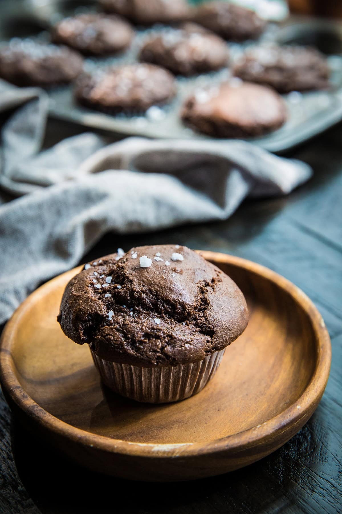 Chocolate zucchini muffin on a wooden plate with the tray of the rest of the muffins in the background.