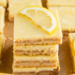 Stack of almond flour lemon bars on a sheet of parchment paper with the rest of the lemon bars all around. A slice of lemon on top of the bars.