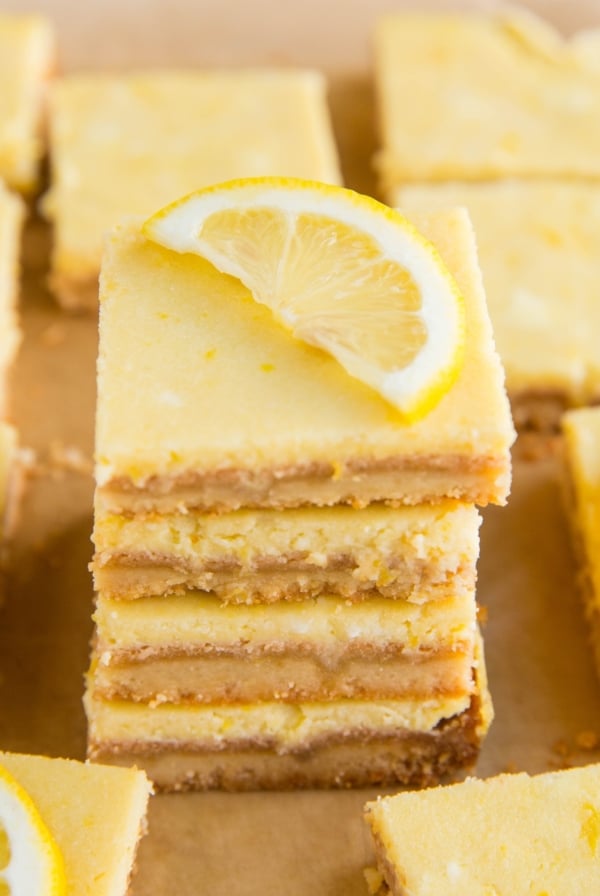 Stack of almond flour lemon bars on a sheet of parchment paper with the rest of the lemon bars all around. A slice of lemon on top of the bars.