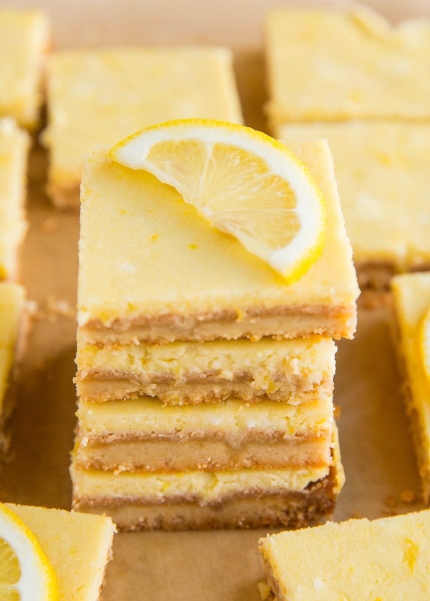 Stack of almond flour lemon bars on a sheet of parchment paper with the rest of the lemon bars all around. A slice of lemon on top of the bars.