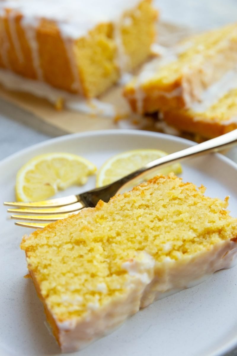 Slice of lemon pound cake on a white plate the fresh lemon slices and a golden fork.