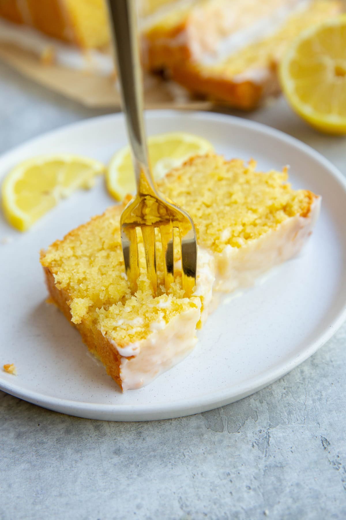 Slice of lemon pound cake on a white plate with a gold fork taking a bite out.