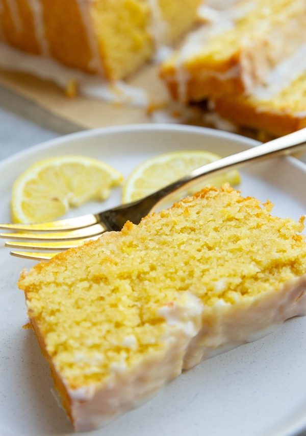 Thick slice of glazed lemon pound cake on a plate with a golden fork and the rest of the loaf in the background.