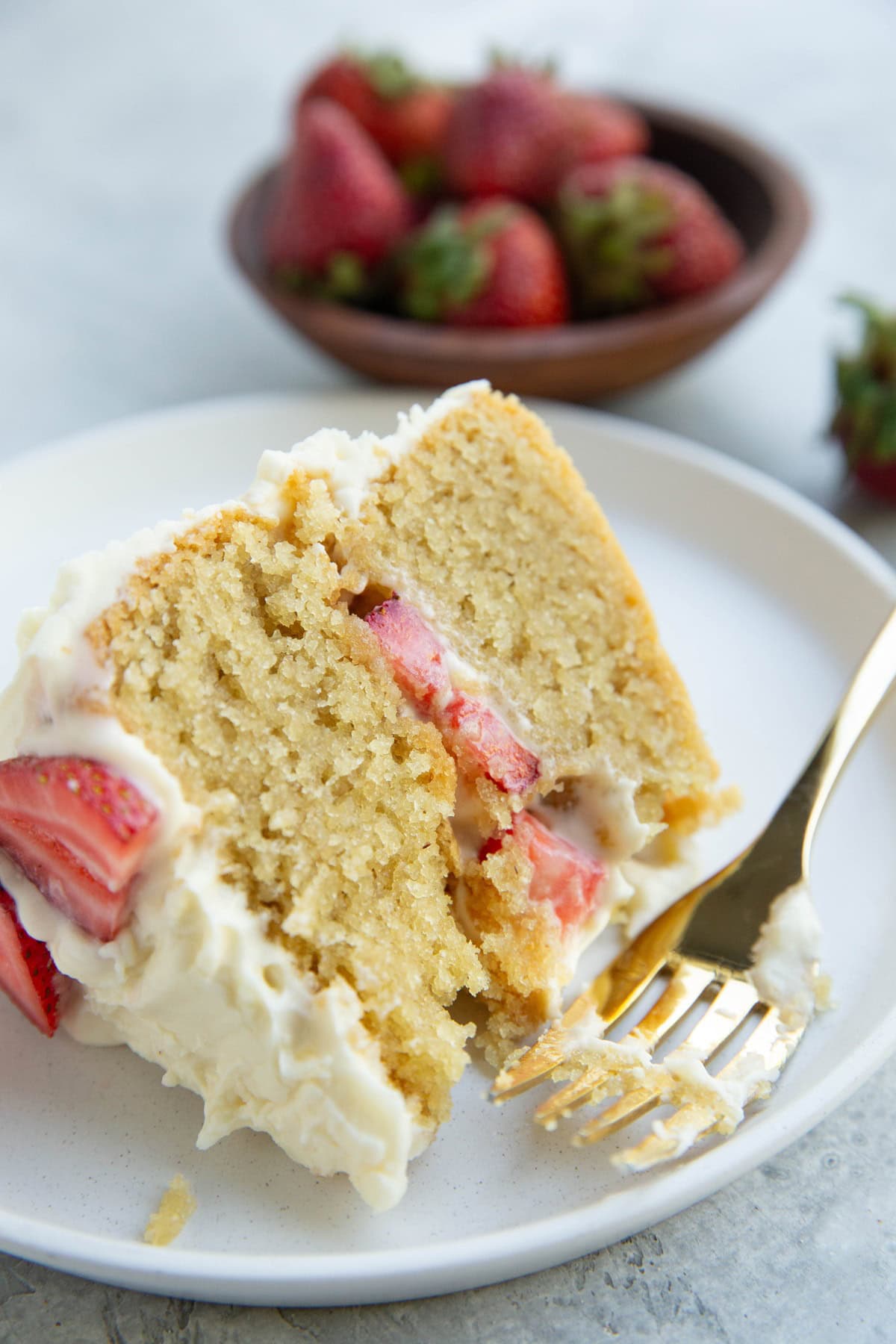 Slice of vanilla layer cake with a bite taken out and a bowl of fresh strawberries in the background.