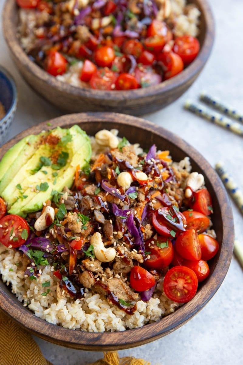 Two large wooden bowls filled with ground turkey mixed with vegetables and sauce on top of cooked brown rice with fresh tomatoes, avocado and cashews. Chop sticks to the side, ready to eat.