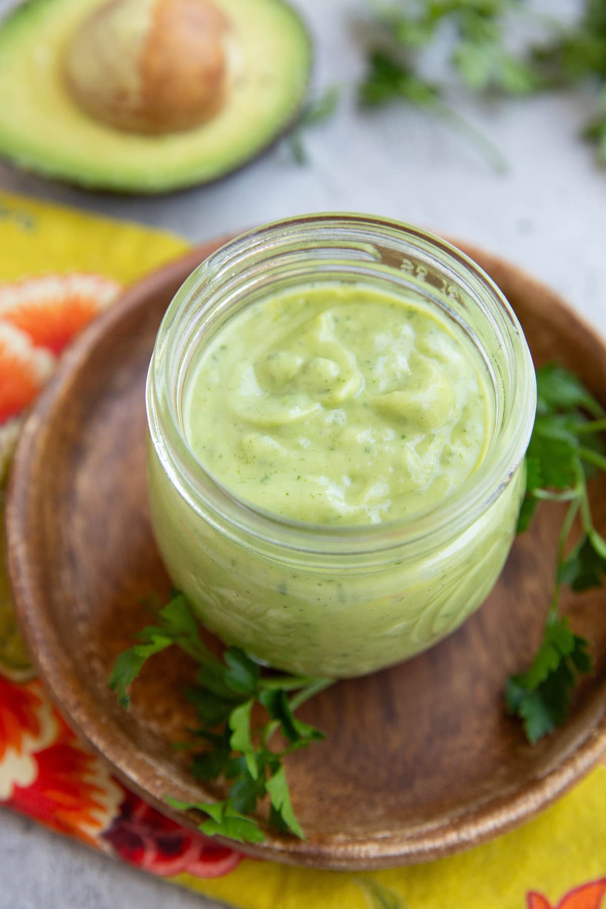 Avocado dressing in a small jar on top of a wooden plate with fresh parsley to the side and an avocado in the background.