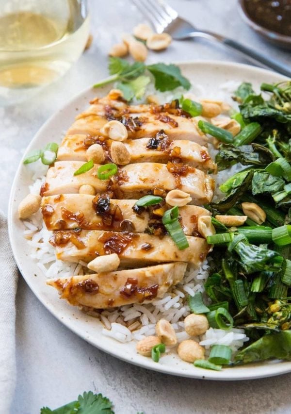 Lemongrass chicken sliced on a plate with white rice and broccolini. A glass of white wine in the background.