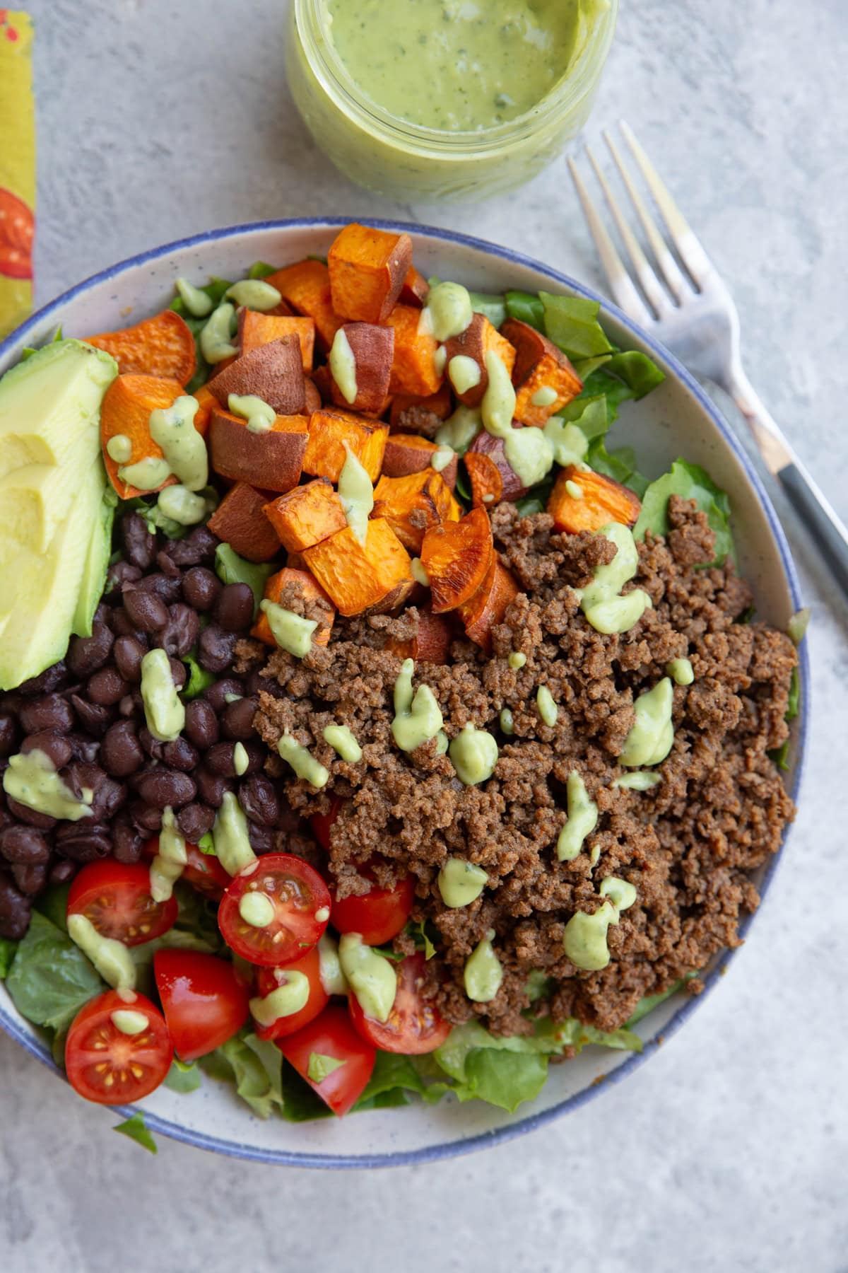 large bowl of ground beef over a bed of chopped greens with rice, roasted sweet potatoes, cherry tomatoes, and drizzled with avocado dressing. A fork to the side.