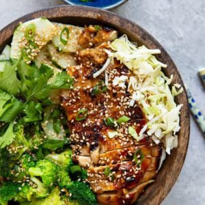 Korean BBQ chicken in a wooden bowl with rice, broccoli, shredded cabbage, cucumber salad, sprinkled with sesame seeds and green onions.