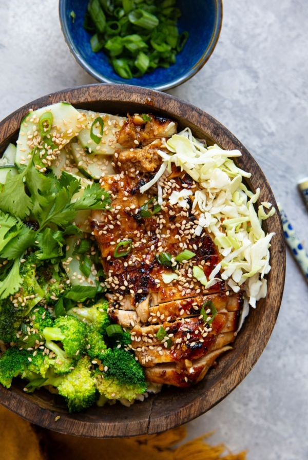 Korean BBQ chicken in a wooden bowl with rice, broccoli, shredded cabbage, cucumber salad, sprinkled with sesame seeds and green onions.