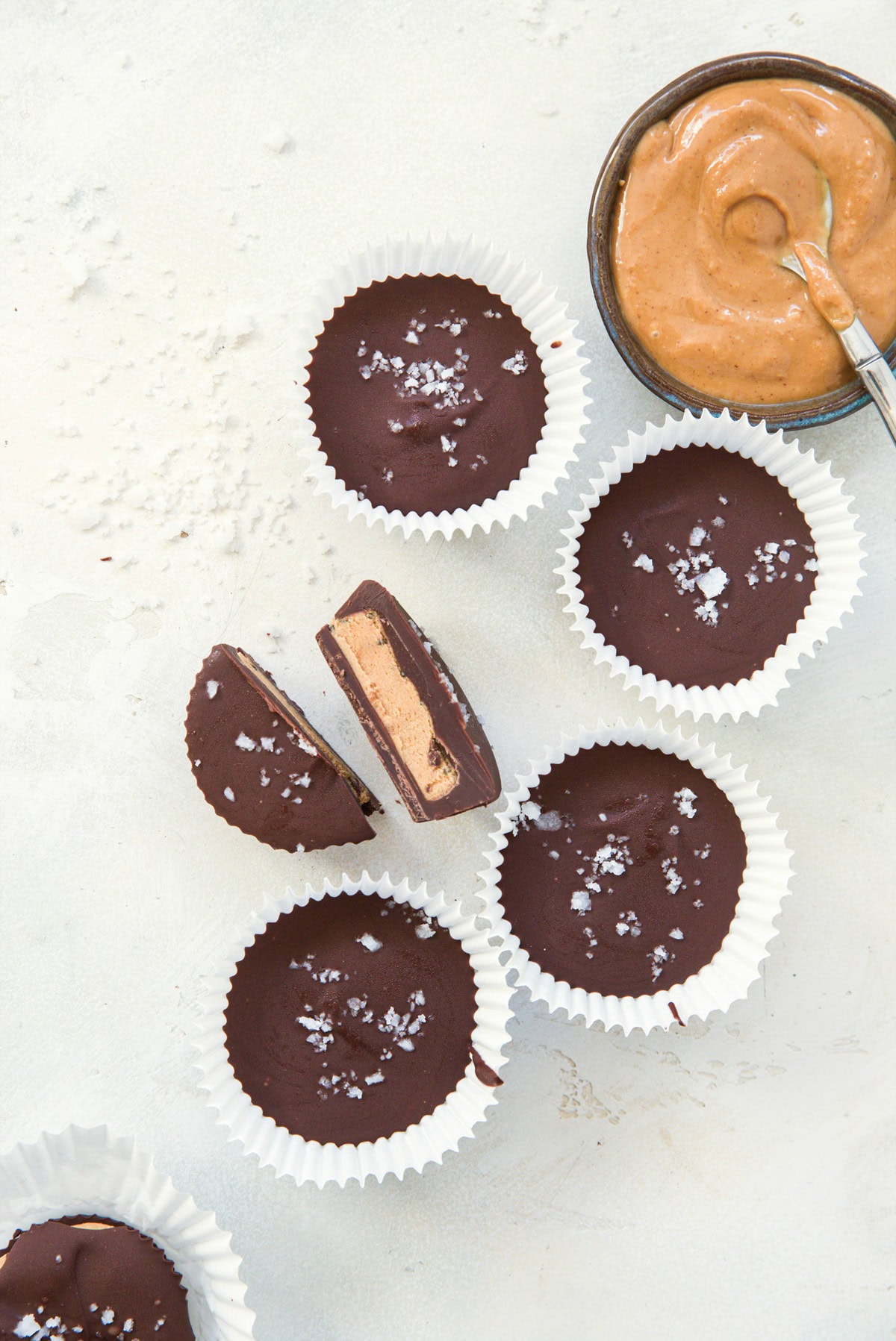 6 peanut butter cups sitting on a white background with one sliced in half, revealing the peanut butter center. a small bowl of peanut butter to the side.