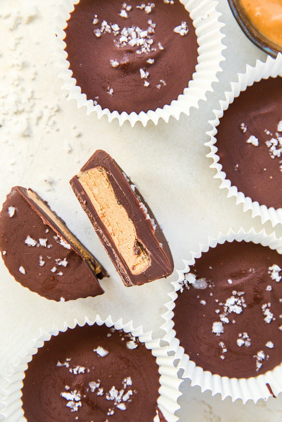 Peanut butter cups sitting on a white background with one sliced in half to reveal the peanut butter center. All cups sprinkled with sea salt.