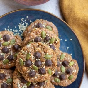 Blue plate of breakfast cookies topped with chocolate chips, pumpkin seeds, and hemp seeds. A bowl of hemp seeds and a golden napkin to the side.
