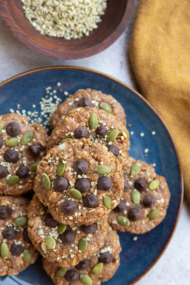 Blue plate of breakfast cookies topped with chocolate chips, pumpkin seeds, and hemp seeds. A bowl of hemp seeds and a golden napkin to the side.