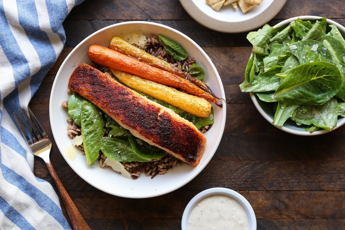 Bowl full of baked salmon, Caesar salad, and roasted carrots. A bowl full of salad to the side, a napkin and a fork, ready to eat.