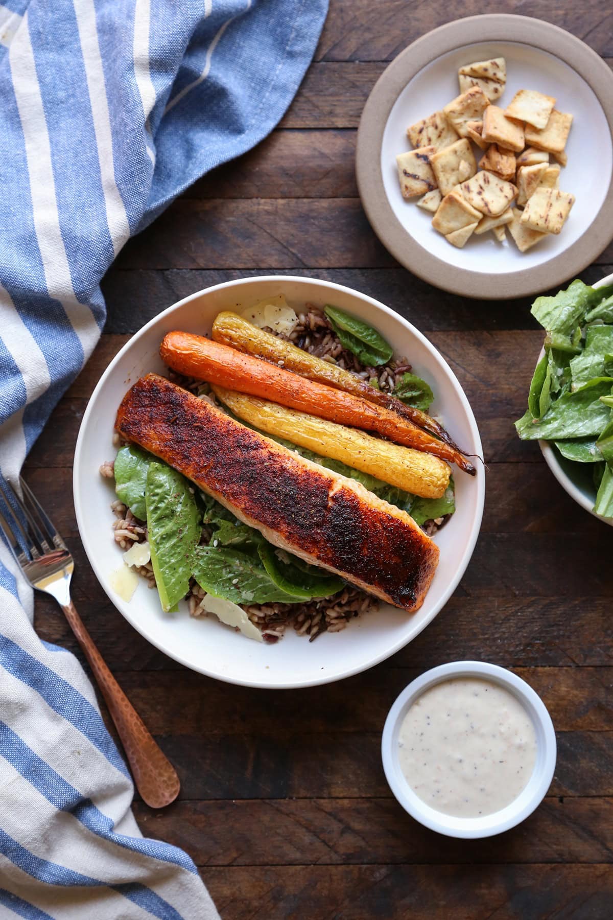 Bowl full of baked salmon, Caesar salad, and roasted carrots with Caesar dressing and more salad to the side.