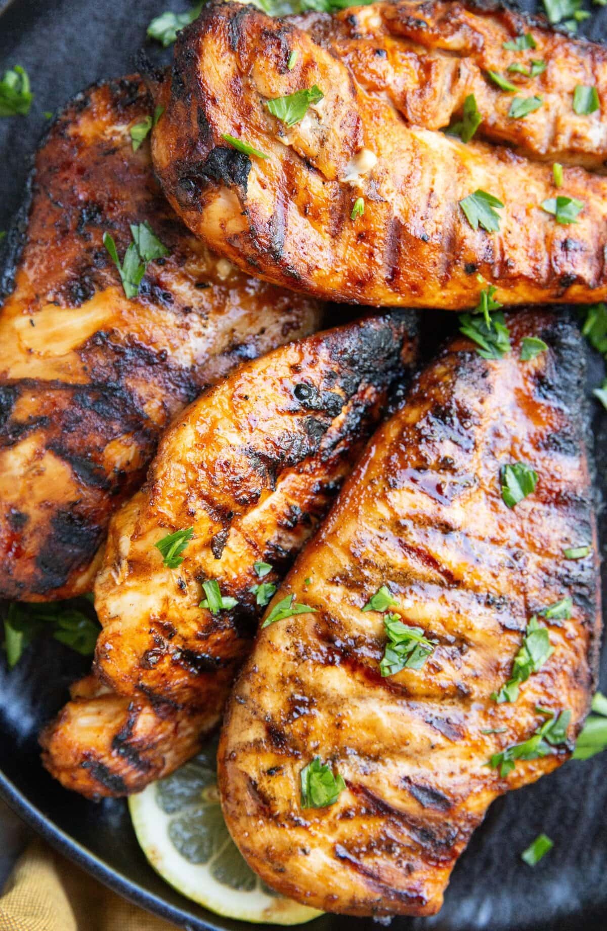Four tequila lime chicken breasts on a black plate with a sprinkle of parsley and lemon slice.
